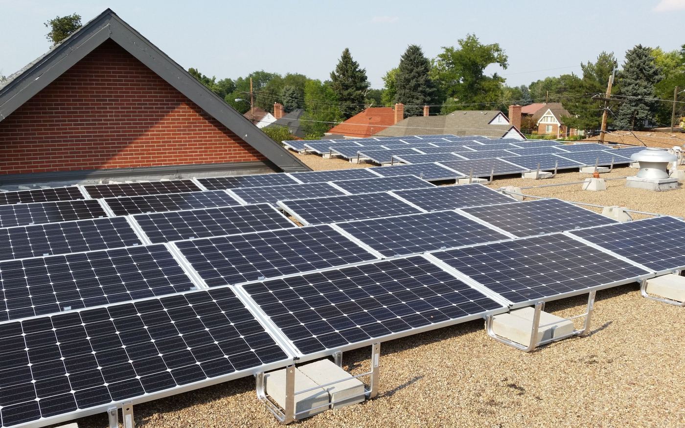 Rows of solar panels on ballasted racks on a flat gravel roof, with a brick gable and a neighborhood behind